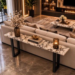 Peut inclure: Une console longue et étroite avec un plateau en pierre à motifs blancs et bruns et deux pieds en métal noir en forme de U. La table supporte un vase en verre brun avec des fleurs blanches, un bol doré sur deux livres et un bougeoir en verre brun.