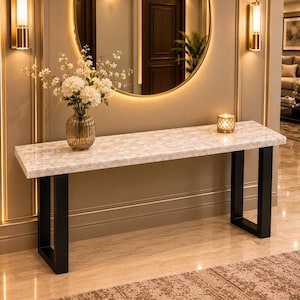 May include: A rectangular console table featuring a white and beige marble-patterned top supported by two black metal U-shaped legs. A clear glass vase filled with white flowers and a small gold-toned candle holder sit on the table surface.
