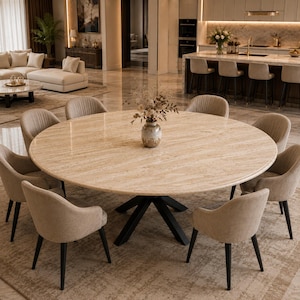 Puede incluir: Una gran mesa de comedor redonda con tablero de piedra travertino beige y base de metal negro, rodeada por ocho sillas tapizadas en tela marrón claro neutra. Un pequeño jarrón de cerámica con flores secas se sitúa en el centro.