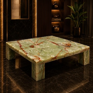 Puede incluir: Una mesa de centro cuadrada hecha de piedra de ónix verde pulido con vetas de color óxido en toda la superficie y cuatro patas gruesas en forma de bloque. Un pequeño vaso transparente con un líquido de color ámbar descansa sobre la mesa.