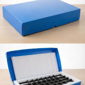 May include: A rectangular blue cardboard box shown closed and open. The interior features a white foam lining and a black grid-like insert for organisation. The text PACKINGS BOX is printed in white on a black background at the bottom right corner.