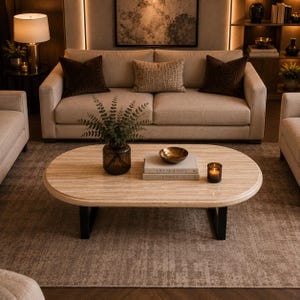 May include: An oval coffee table with a light beige travertine stone top and black metal legs. On the table sits a dark glass vase with green foliage, a small gold bowl, a lit candle in a glass holder, and two books titled Live Beautiful and Kinfolk.