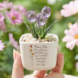 May include: A small white ceramic pot with a glass flower arrangement. The pot has a floral design and text that reads "Bonus Mom". The glass flowers are purple and the leaves are green. The pot is filled with small white stones.