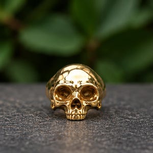 May include: A polished gold-tone ring shaped like a human skull. The ring features a smooth, reflective surface with detailed eye sockets, a nasal cavity, and defined teeth, showcasing a metallic finish that catches the light.