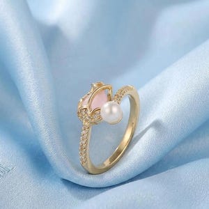 May include: A gold-colored ring featuring a tulip design with a pink center and a white pearl. The ring is encrusted with small, clear gemstones along the band. The ring is set against a light blue fabric background.