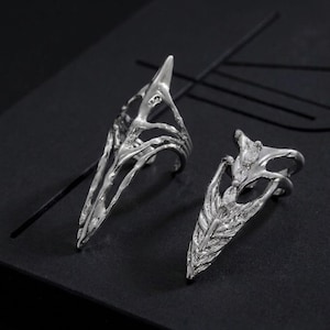 May include: Two silver-colored finger rings with a gothic, claw-like design. The rings have a sharp, pointed tip and intricate openwork detailing, creating a unique and edgy aesthetic. The rings are displayed on a dark surface.