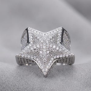 May include: A silver-toned star-shaped ring, encrusted with numerous small, clear gemstones. The ring's design features a five-pointed star, with the gemstones covering the entire surface, creating a sparkling effect. The ring is set against a soft gray background.