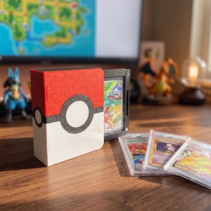 Pode incluir: Um porta-cartões vermelho, branco e preto projetado para se assemelhar a uma Pokébola. O suporte está ao lado de uma vitrine de cartas preta e vários cartões colecionáveis classificados. Uma figura azul e branca está no fundo.