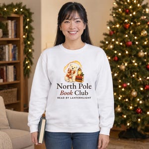 May include: A white long-sleeve sweatshirt featuring a graphic of a polar bear wearing a red and white striped scarf next to a lantern and books. The text reads North Pole Book Club and READ BY LANTERNLIGHT in black and red lettering.