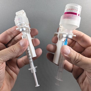 May include: Two white plastic medical vial adapters attached to glass vials and syringes. The left syringe is marked with 0.1 to 1.0 ml increments. The right syringe is marked with 1.5 to 3 ml increments. The vials feature white caps and labels.