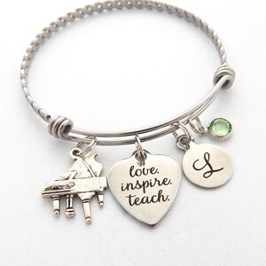 Peut inclure: Un bracelet &agrave; breloques en argent avec un charme de piano, un charme en forme de c&oelig;ur avec les mots "love, inspire, teach", un charme rond avec la lettre "L" et un charme en cristal vert.