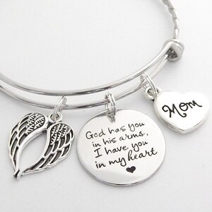 May include: A silver expandable bracelet with three charms. The charms are a pair of angel wings, a round charm with the text "God has you in his arms, I have you in my heart", and a heart charm with the word "Mom" engraved on it.