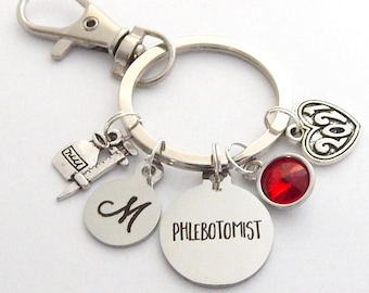 May include: Silver keychain with a red rhinestone, a charm with the word "PHLEBOTOMIST", a charm with the letter "M", a charm with a syringe, and a heart charm with the number "2021".
