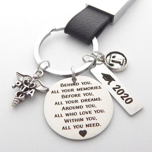 May include: Silver keyring with a black leather strap, a round charm with the text "BEHIND YOU, ALL YOUR MEMORIES. BEFORE YOU, ALL YOUR DREAMS. AROUND YOU, ALL WHO LOVE YOU. WITHIN YOU, ALL YOU NEED." and a heart charm. There are also charms with the text "2020", a graduation cap, and a medical symbol.