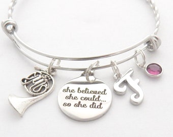 Puede incluir: Un brazalete de plata con un dije que dice "ella creía que podía... así que lo hizo." El brazalete también tiene un dije de trompa francesa, un dije de la letra "J" y una pequeña piedra preciosa púrpura.
