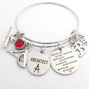 May include: A silver charm bracelet with a red crystal charm, a graduation cap charm, a protractor charm, a charm with the word "ARCHITECT" and a compass symbol, and a charm with the letter "B". The bracelet also has a charm with the text "Behind you. all your memories. Before you. all your dreams. Around you. all who love you. Within you. all you need."