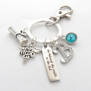 May include: Silver keychain with a graduation scroll charm, a medical symbol charm, a charm with the text "she believed she could... so she did", a rhinestone letter B charm, and a light blue rhinestone charm.