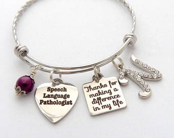 May include: Silver charm bracelet with a purple bead, a heart charm with the text "Speech Language Pathologist", a square charm with the text "Thanks for making a difference in my life", and a letter "M" charm with rhinestones.