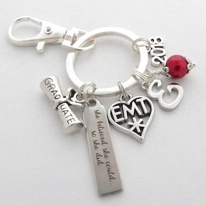 May include: Silver keyring with charms including a graduation scroll, a heart with the letters EMT, a charm with the number 3, a red bead, and a tag with the text "she believed she could... so she did".