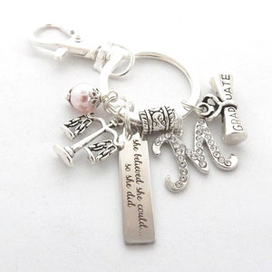 May include: Silver keychain with charms, including a graduation cap, a letter M with rhinestones, a scale of justice, a pink pearl, and a tag with the text "she believed she could... so she did."