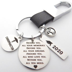 May include: A silver keyring with a black leather strap and a large silver clasp. The keyring has several charms, including a round charm with the word "CHIROPRACTOR" and a graduation cap charm with "2020". A round charm has an engraved message.