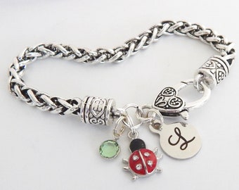Puede incluir: Pulsera de plata con un dije de mariquita, una piedra preciosa verde, un dije de corazón y un dije de plata con la letra "L" grabada.