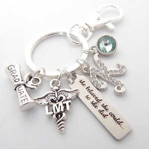 May include: Silver keychain with charms, including a graduation cap, a medical caduceus with the letters LMT, a charm with the text "she believed she could... so she did", and a silver letter K with rhinestones. The keychain also has a light blue crystal charm.