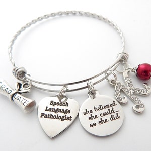 May include: Silver charm bracelet with a graduation cap, a heart charm that says "Speech Language Pathologist", a round charm that says "She believed she could... so she did", a letter K charm with rhinestones, and a red bead.