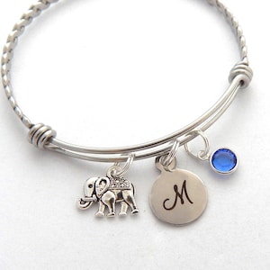 May include: A silver expandable bangle bracelet with a charm of a silver elephant, a silver charm with the letter "M" engraved on it, and a blue crystal charm.
