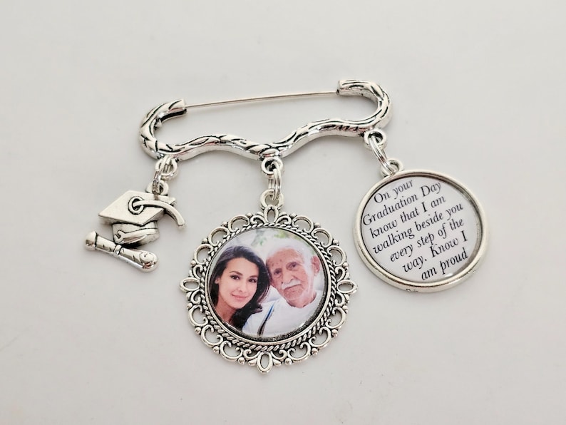May include: Silver graduation pin with a photo charm, a graduation cap charm, and a charm with the text "On your Graduation Day know that I am walking beside you every step of the way. Know I am proud."