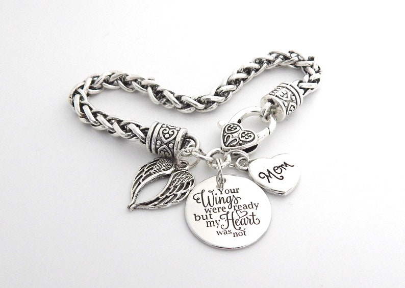 Op de afbeelding: Zilveren armband met een bedel met de tekst "Your wings were ready but my heart was not". De armband heeft ook een hartbedel met de tekst "Mom" en een kleine engelenvleugelbedel.