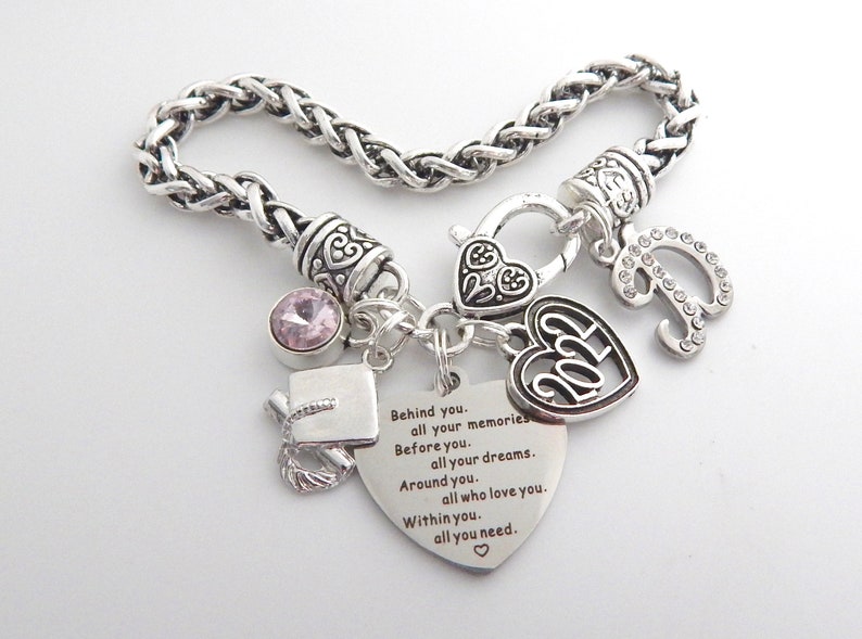 K&ouml;nnte beinhalten: Ein silbernes Armband mit einem Abschluss-Hut-Anh&auml;nger, einem Herz-Anh&auml;nger mit der Jahreszahl 2024, einem Herz-Anh&auml;nger mit einer Inschrift, einem rosa Kristall-Anh&auml;nger und einem Buchstaben-P-Anh&auml;nger mit Strasssteinen.