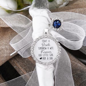 May include: A silver brooch pin featuring a round pendant with a scalloped rhinestone border and the text: TODAY A BRIDE TOMORROW A WIFE FOREVER OUR LITTLE GIRL MOM & DAD. The pin is adorned with a white ribbon and a square blue rhinestone accent.