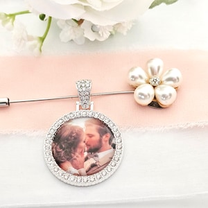 Könnte beinhalten: Silberfarbene Anstecknadel mit rundem Anhänger, der ein Foto eines sich küssenden Paares zeigt, umgeben von klaren Kristallen. Eine Perlenblumen-Anstecknadel ist ebenfalls vorhanden, Hochzeitsthema.