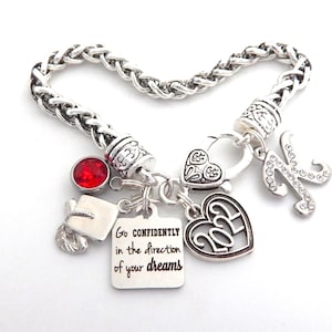 K&ouml;nnte beinhalten: Silbernes Armband mit einem roten Strassstein, einem Abschluss-Hut-Anh&auml;nger, einem Herz-Anh&auml;nger mit dem Jahr 2023, einem Herz-Anh&auml;nger mit einem Design, einem Buchstaben K-Anh&auml;nger mit Strasssteinen und einem Anh&auml;nger mit dem Text "Go confidently in the direction of your dreams".