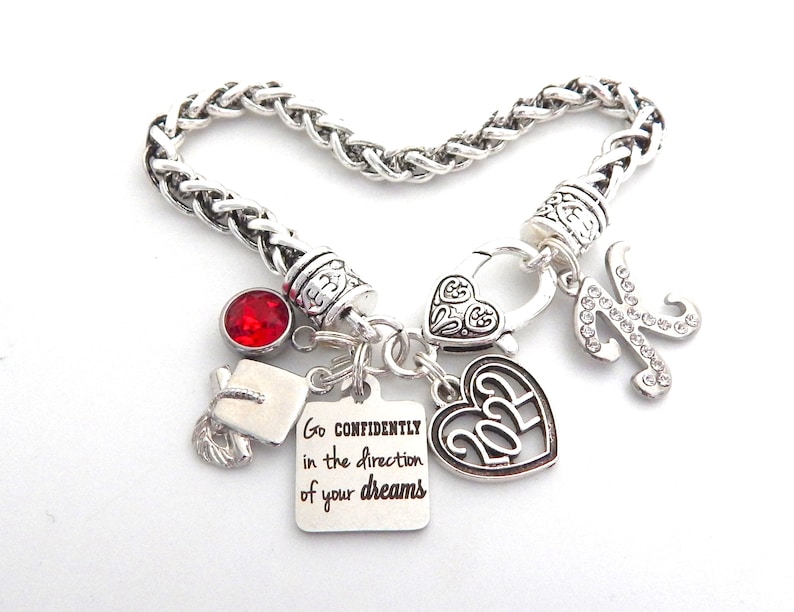 K&ouml;nnte beinhalten: Silbernes Armband mit einem roten Strassstein, einem Abschluss-Hut-Anh&auml;nger, einem Herz-Anh&auml;nger mit dem Jahr 2023, einem Herz-Anh&auml;nger mit einem Design, einem Buchstaben K-Anh&auml;nger mit Strasssteinen und einem Anh&auml;nger mit dem Text "Go confidently in the direction of your dreams".