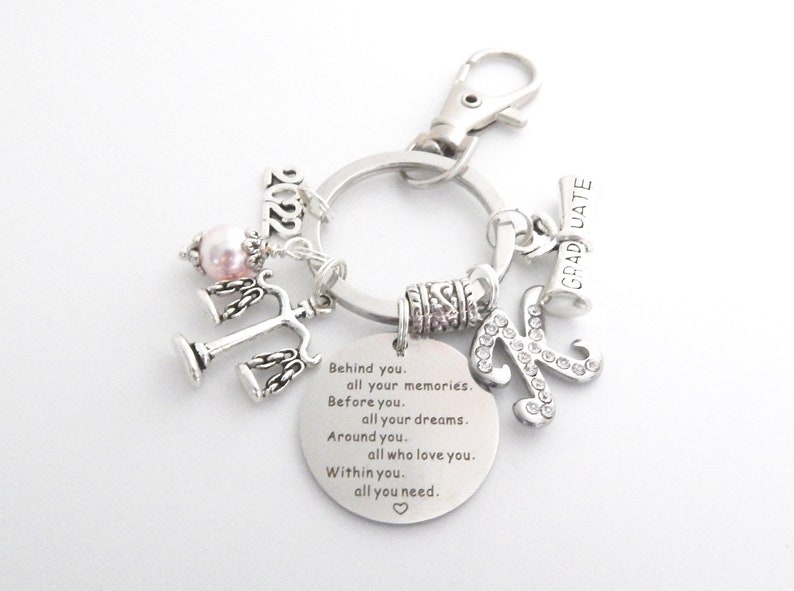 May include: Silver keychain with a charm that says "GRADUATE" and a charm with a letter K covered in rhinestones. The keychain also has a charm with a scale of justice, a pink pearl, and a charm with the year 2022. The keychain has a round tag with the text "Behind you. all your memories. Before you. all your dreams. Around you. all who love you. Within you. all you need."
