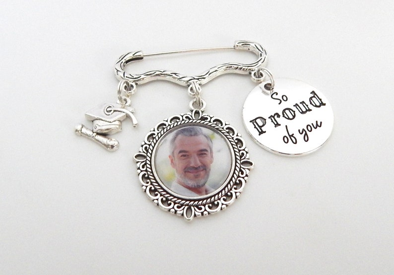 May include: A silver graduation pin with a photo charm, a graduation cap charm, and a round charm that says 'So Proud of you'.