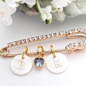 May include: A gold safety pin with a row of clear rhinestones. The pin has three charms attached to it: a white mother-of-pearl disc with the letter "B" engraved on it, a blue heart charm, and a white mother-of-pearl disc with the letter "K" engraved on it.