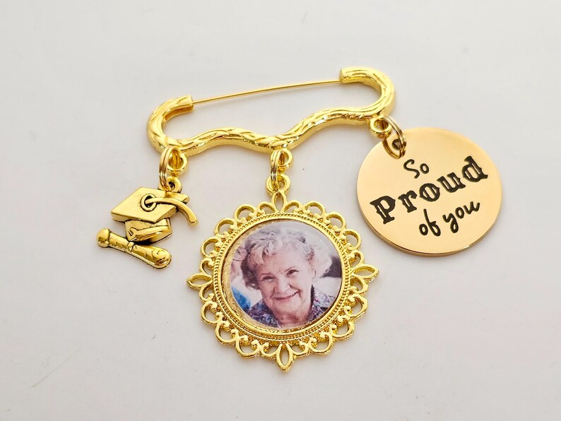 May include: Gold graduation pin with a photo charm, a graduation cap charm, and a round charm that says "So Proud of You".
