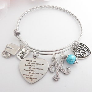 K&ouml;nnte beinhalten: Ein silbernes Armband mit Charms, darunter eine Abschluss-Kappe, ein Herz-Anh&auml;nger mit dem Text "You are loved!" und ein Herz-Anh&auml;nger mit dem Text "Behind you. all your memories. Before you. all your dreams. Around you. all who love you. Within you. all you need." Das Armband hat au&szlig;erdem einen Anh&auml;nger mit dem Buchstaben "K" und eine t&uuml;rkisfarbene Perle.