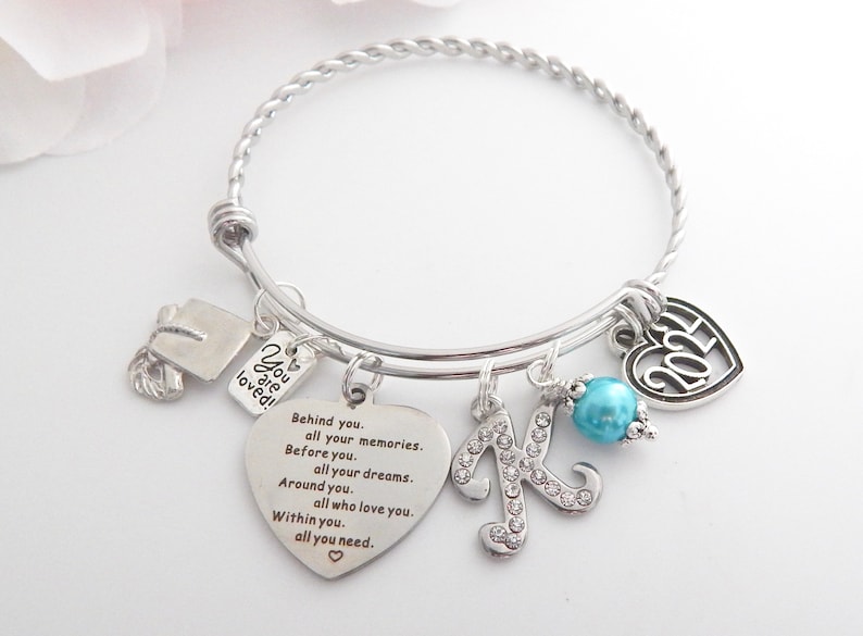 K&ouml;nnte beinhalten: Ein silbernes Armband mit Charms, darunter eine Abschluss-Kappe, ein Herz-Anh&auml;nger mit dem Text "You are loved!" und ein Herz-Anh&auml;nger mit dem Text "Behind you. all your memories. Before you. all your dreams. Around you. all who love you. Within you. all you need." Das Armband hat au&szlig;erdem einen Anh&auml;nger mit dem Buchstaben "K" und eine t&uuml;rkisfarbene Perle.