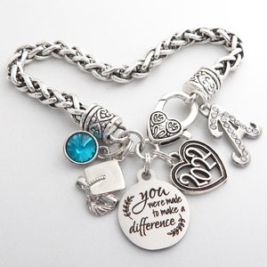 K&ouml;nnte beinhalten: Silbernes Armband mit Charms: ein Abschluss-Hut, ein blauer Kristall, ein Herz-Anh&auml;nger mit dem Jahr 2023, ein Buchstabe A und ein runder Anh&auml;nger mit dem Text "You were made to make a difference".