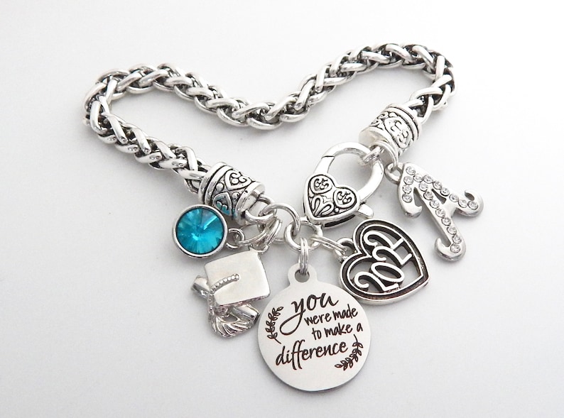 K&ouml;nnte beinhalten: Silbernes Armband mit Charms: ein Abschluss-Hut, ein blauer Kristall, ein Herz-Anh&auml;nger mit dem Jahr 2023, ein Buchstabe A und ein runder Anh&auml;nger mit dem Text "You were made to make a difference".