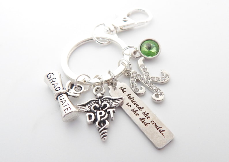 May include: Silver keychain with charms including a graduation cap, a medical symbol with "DPT" engraved, a charm with the text "she believed she could... so she did", a green rhinestone, and a silver letter K with rhinestones.
