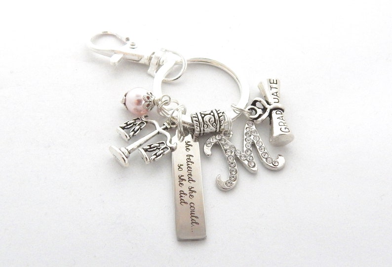 May include: Silver keychain with charms, including a graduation cap, a letter M with rhinestones, a scale of justice, a pink pearl, and a tag with the text "she believed she could... so she did."