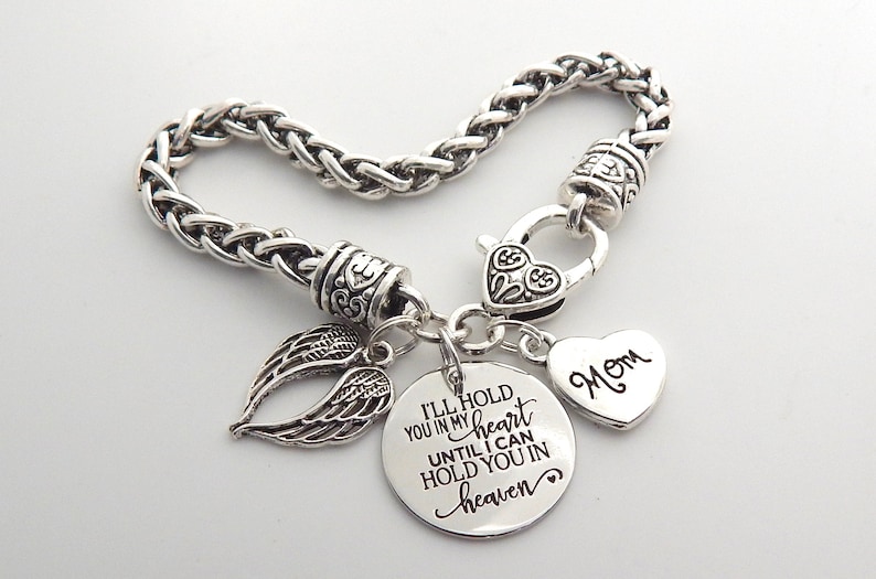 Op de afbeelding: Zilveren armband met een hartvormige bedel met de tekst "Mom" en een ronde bedel met de tekst "Ik zal je in mijn hart houden totdat ik je in de hemel kan vasthouden". De armband heeft ook een kleine engelenvleugelbedel.