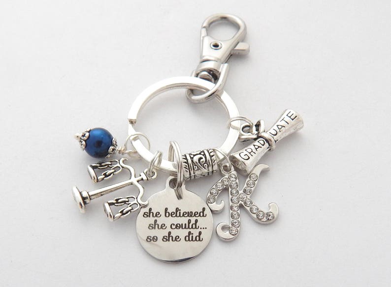 May include: Silver keychain with charms including a graduation cap, a scale of justice, a blue bead, and a letter K. The keychain also has a silver charm with the text "she believed she could... so she did."