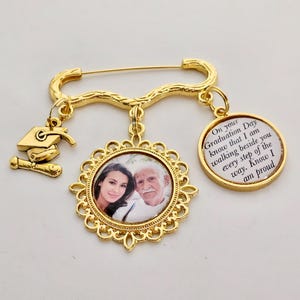 May include: Gold-toned graduation pin with a photo charm of a man and woman, a graduation cap charm, and a charm with the text "On your Graduation Day know that I am walking beside you every step of the way. Know I am proud."