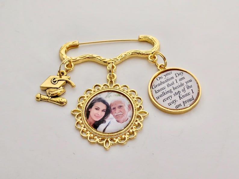 May include: Gold-toned graduation pin with a photo charm of a man and woman, a graduation cap charm, and a charm with the text "On your Graduation Day know that I am walking beside you every step of the way. Know I am proud."